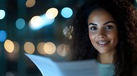 Young African American woman with curly hair smiling while reading document in evening with blurred bokeh lights background, expressing joy and satisfaction.