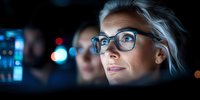 Professional woman with glasses illuminated by computer screen light working late at night in dark office, focused expression while analyzing data and digital information.