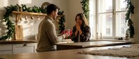 Young couple sharing warm moment in festive kitchen during Christmas morning, man offering gift to smiling woman by decorated fireplace with garland.