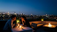 Young Caucasian couple enjoying a romantic candlelit rooftop dinner with a glowing city skyline at night, fire pit nearby.