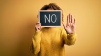 Young man in yellow sweater holding chalkboard with NO text and showing stop hand gesture against warm background for rejection and refusal concepts.