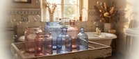 Collection of vintage colored glass jars and bottles arranged on wooden tray in bright kitchen interior with natural sunlight and floating soap bubbles.