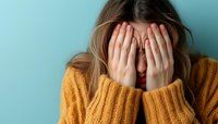Young Caucasian woman in yellow sweater covering face with hands against blue wall, expressing emotional distress, anxiety, shame or sadness.