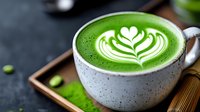 Green matcha latte with white foam art heart pattern in ceramic cup on wooden tray, scattered matcha powder and fresh leaves on dark background.