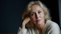 Thoughtful mature blonde woman in cream sweater resting chin on hand with contemplative expression against dark background for lifestyle content.