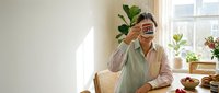 Young woman sitting at bright kitchen table, holding coffee mug covering her face, cozy morning home atmosphere with natural light and plants.