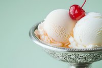 Vanilla ice cream scoops topped with a red cherry in an ornate silver bowl on a soft mint green background.