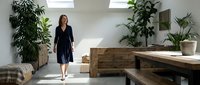 Woman in dark navy wrap dress standing in modern office space with wooden desk, potted plants, and natural light from windows, suitable for corporate, business, and workplace design projects.