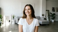 Young brunette woman smiling in modern minimalist living room with white shirt, natural lighting and contemporary home interior design for lifestyle content.