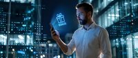 Man holding smartphone with glowing gift box hologram icon in night cityscape. Caucasian male professional using mobile technology for online shopping and digital commerce applications.