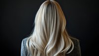 Blonde woman with long wavy hair shown from behind against dark background, wearing gray sweater. Professional hairstyling and hair care concept.