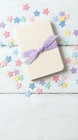 Cream gift box with purple polka dot ribbon surrounded by colorful paper stars on white wooden background for celebrations and special occasions.