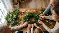 Senior Caucasian man and young woman crafting Christmas wreath together at wooden table with festive decorations and warm lighting in cozy home interior.