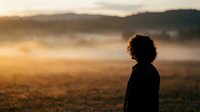 Silhouette of young woman with curly hair standing in misty field during golden hour sunrise. Peaceful morning landscape with rolling hills and warm sunlight.