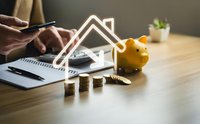 Person calculating home savings with piggy bank, house model and coins on wooden desk for mortgage planning and real estate investment budgeting.