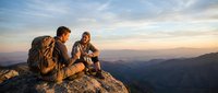 Two hikers with backpacks sitting on mountain summit during golden hour sunset, enjoying panoramic valley views after successful trek.