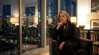 Middle-aged woman with blonde hair, Caucasian ethnicity, wearing dark business attire, sitting thoughtfully in modern apartment with city skyline view at night, suitable for corporate, lifestyle, and professional concepts.