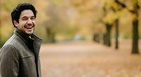 Young Middle Eastern man with dark curly hair smiling at camera in autumn park setting with golden foliage and tree-lined path, wearing casual gray jacket for lifestyle and portrait use.