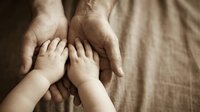 Adult hands gently holding small child feet in warm sepia tone, representing family bond, protection, care, love and parenting connection on textured fabric.