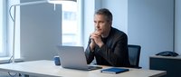 Thoughtful mature businessman in suit contemplating strategy while working on laptop in modern office environment with natural lighting.