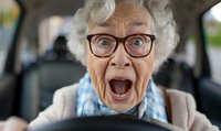 Senior woman with gray curly hair and glasses expressing extreme surprise while driving car, mouth wide open in shock or excitement, wearing cardigan.