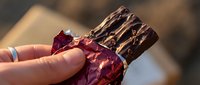 Unrecognizable person holding chocolate-covered confection with red foil wrapper, close-up detail shot with shallow depth of field and warm natural lighting.