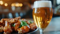 Cold glass of light beer with foam head next to spicy buffalo chicken wings garnished with fresh herbs, bokeh lights in background for restaurant menu.