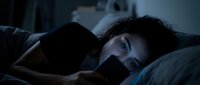 Young woman with dark hair and fair complexion lying in bed at night, looking at smartphone screen with concerned expression, experiencing insomnia or late-night anxiety.