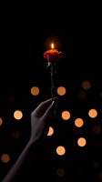 Female hand holding red rose with burning candle on top against dark background with golden bokeh lights creating magical romantic atmosphere.