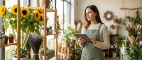 Young Caucasian female florist in green apron using tablet in bright flower shop surrounded by sunflowers and plants for small business management.