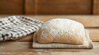 Fresh raw bread dough sprinkled with flour on white cloth and rustic wooden table with checkered kitchen towel in background, ready for baking.