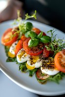 Fresh caprese salad with sliced mozzarella, ripe tomatoes, basil leaves and microgreens on white plate, garnished with black pepper and olive oil. Selective focus.