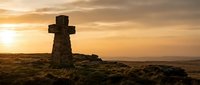 Stone cross monument standing on moorland at golden hour sunset, casting long shadows across rugged terrain. Religious landmark.