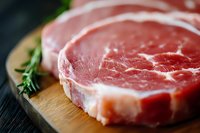 Fresh raw ribeye steak with marbling and fat cap on wooden cutting board with rosemary sprig, macro close-up view showing meat texture and quality.