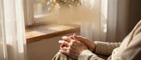Elderly woman hands clasped in prayer by sunny window with warm golden light streaming through white curtains creating peaceful spiritual moment.