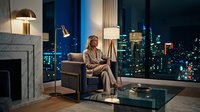 Woman in business casual attire sitting in modern luxury apartment with floor-to-ceiling windows overlooking illuminated city skyline at night, contemporary interior design with fireplace and elegant furnishings.