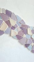 Colorful sea glass pieces in purple, lavender, cream and blue tones scattered on white textured surface for craft projects and coastal home decor.