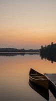 Wooden boat moored at dock on calm lake during golden sunrise, with forested shoreline reflected in still water. Suitable for travel, nature, and relaxation themes.