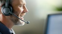 Smiling mature bearded man wearing headset while working at computer in office environment. Professional customer service representative providing remote support.