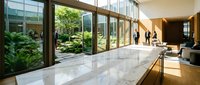 Modern office lobby with floor-to-ceiling windows overlooking landscaped courtyard garden, polished marble floors and contemporary seating area.