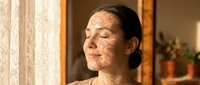 Middle-aged woman with East Asian ethnicity applying facial mask treatment at home near window with natural light and warm interior decor.