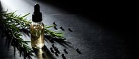 Natural rosemary essential oil in glass dropper bottle with fresh green herbs and scattered seeds on dark stone surface for aromatherapy and wellness.