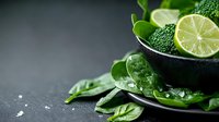 Young woman with light skin tone, wearing professional chef attire, preparing fresh green ingredients in mortar and pestle. Lime slices, spinach leaves, and herbs arranged on dark surface for culinary or wellness content.