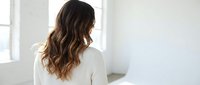 Young woman with long wavy brown hair wearing white sweater sitting on bed in bright minimalist bedroom with natural light from window.