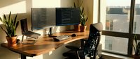 Modern home office workspace with dual monitors displaying code on wooden desk near window with natural lighting and plants for productivity.
