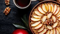 Fresh apple oatmeal topped with sliced red apples and walnuts served with hot tea on dark background, overhead view of healthy breakfast bowl.