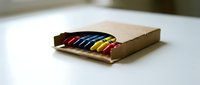 Colorful wax crayons in a cardboard box on a white table, featuring red, yellow, and black colors for art and education.