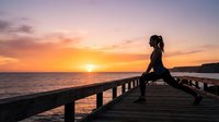 Athletic woman stretching on wooden pier at sunset over ocean. Female athlete performing yoga or fitness routine during golden hour by seaside for wellness and exercise content.