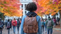 Young African American student with leather backpack walking on college campus during autumn season with vibrant fall foliage and blurred students in background.