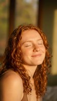 Young redhead woman with curly hair and freckles enjoying warm sunlight with eyes closed in peaceful moment outdoors during golden hour sunset.